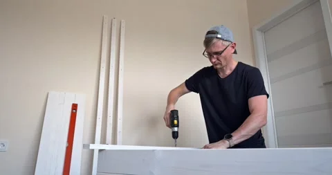 Man assembling white wooden shelving unit indoors using screwdriver to tighten Stock Footage 314789210