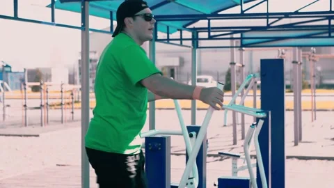 A man athlete in sunglasses does exercises on a machine tool on an outdoor Stock Footage 147925323