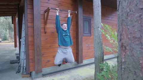 Man Attempting Pull Ups on Outdoor Bar Near Wooden House Stock Footage 334331557