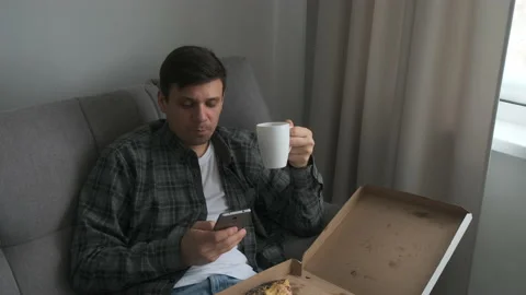 Man bachelor sits on sofa eating tasty pizza, drinking tea, browsing smartphone. Stock Footage 153890766