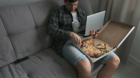 Man bachelor sits on sofa eating tasty pizza, drinking tea, watching in laptop. Stock Footage 159489236