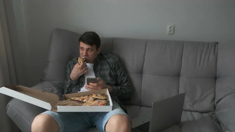 Man bachelor sits on sofa eating tasty pizza, drinking tea, browsing smartphone. Video stock 159743791