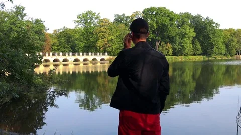 A man (back to the camera) talks on a smartphone - a river in a rural area in Video stock 82241403