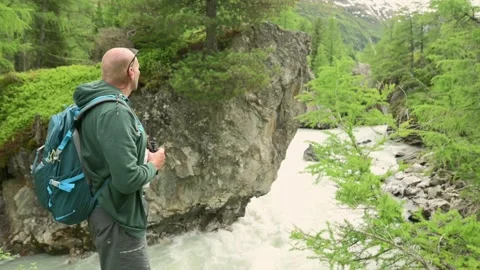 A man with a backpack and binoculars hiking along a river in a scenic mountain Vidéo 281107534