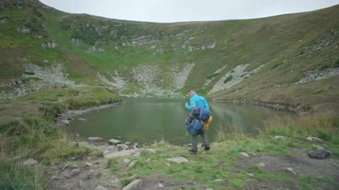 A man with a backpack and a dog approaches a mountain lake Stock Footage 220094791