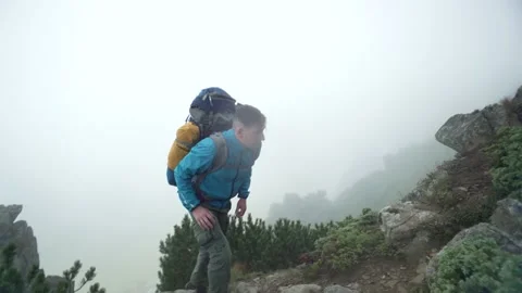 A man with a backpack climbs rocky mountains into the fog. A tourist in the rain Stock Footage 220094860
