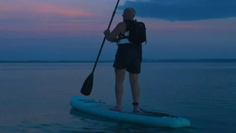 A man with a backpack floats on a SUP board on the smooth water of the lake at Stock Footage 205319810