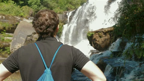 Man with backpack gazes at cascading waterfall in lush green forest, Stock Footage 280020156