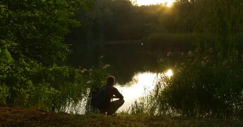 Man With Backpack On His Back Sits On His Haunches At The Lakeshore He Stock Footage 55497920