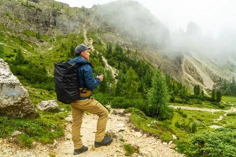Man with backpack holding steaming mug in rainy Dolomite mountains, Italy. Fo Stock Photos