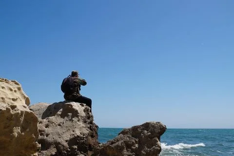A man with a backpack looks through binoculars at the sea horizon while sit.. Stock Photos