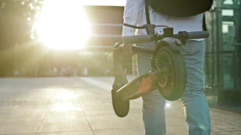 Man with a backpack over his shoulders carries a folded scooter along a city Stock Footage 199218319