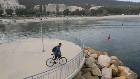 A man with a backpack rides a bicycle along a seafront promenade, seen from a Video stock 255648275
