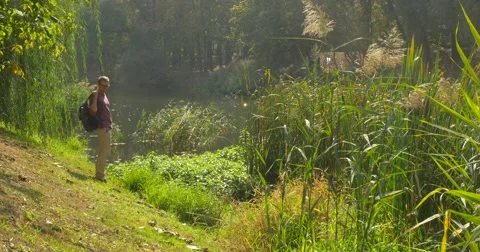 Man With Backpack Sits to The Grass at The Sloping Lake Bank Man is Looking at Stock Footage 56067631