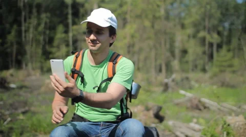 A man with a backpack sits on an old stump of a tree. Clicks on the phone Stock Footage 64429920
