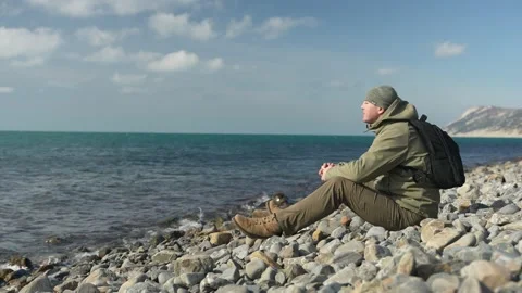A man with a backpack sits on a rocky beach and enjoys a beautiful view of the 動画素材 236569944