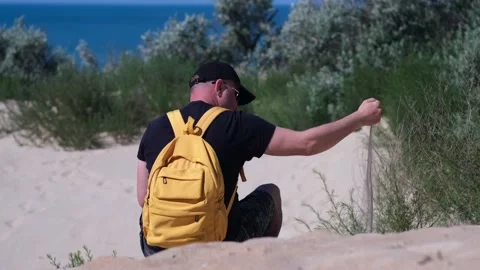 Man with backpack sits on sand beach dune, sand puffs through his fingers Stock-Footage 235906639