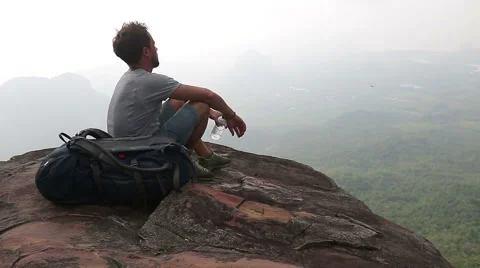 A man with backpack sitting on a rock drinking water with a valley at the bottom 스톡 동영상 49456243
