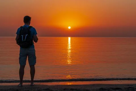A man with a backpack stands on the beach and watches the sun rise. Foto stock