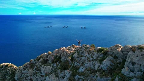 A man with a backpack stands on the edge of a cliff overlooking the ocean. .. Video stock 233335725