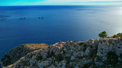 A man with a backpack stands on the edge of a cliff overlooking the ocean. .. Video stock 233543402