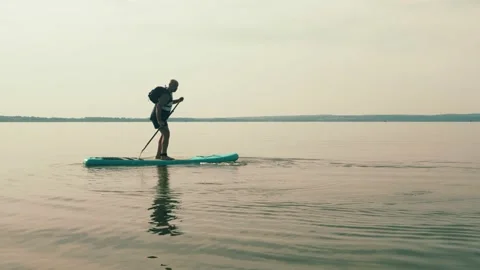 A man with a backpack on a SUP board swims in the lake on a sunny day against Stock Footage 219505036