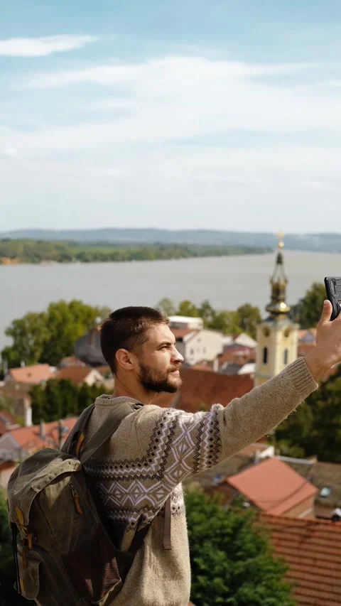 A man with a backpack takes a selfie on a hill with a river, rooftops, and a Stock Footage 318542143