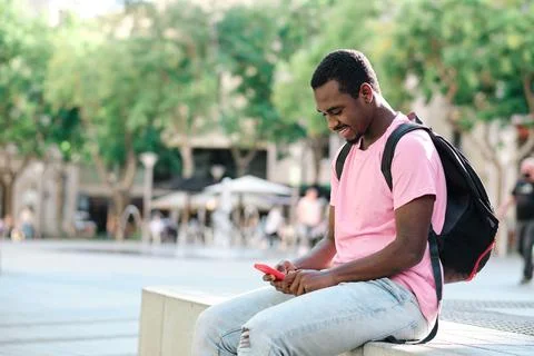 Man with backpack using a mobile phone while sitting on a bench outdoors. Stock Photos