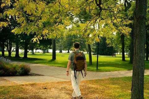 A man with a backpack walks along a path in a lush green park setting. Stock Photos