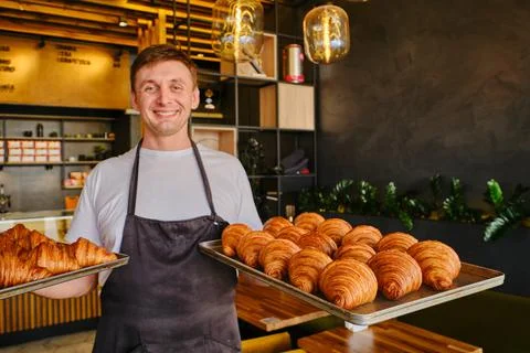 Man baker holds two baking sheets of fresh golden croissants at the bakery Stock Photos