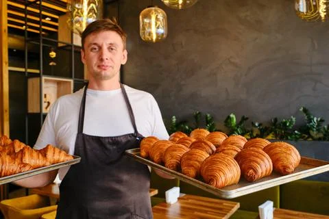 Man baker holds two baking sheets of fresh golden croissants at the bakery Stock Photos