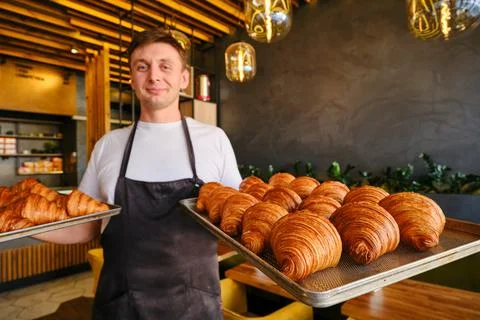 Man baker holds two baking sheets of fresh golden croissants at the bakery Stock Photos