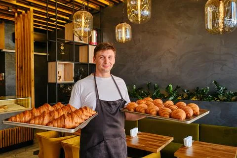 Man baker holds two baking sheets of fresh golden croissants at the bakery Stock Photos