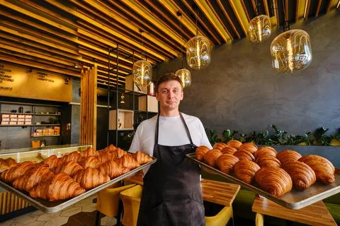 Man baker holds two baking sheets of fresh golden croissants at the bakery Stock Photos
