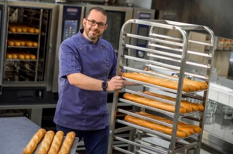 Man baker in uniform baking baguettes with bread shelves on background 스톡 사진