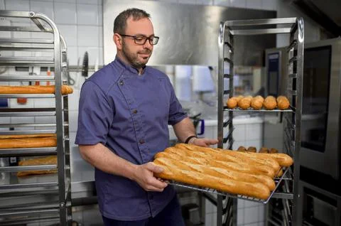 Man baker in uniform holding baguettes on tray with bread shelves on background 스톡 사진
