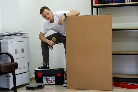Man balanced on toolbox drilling side of cardboard box in room Stock Photos