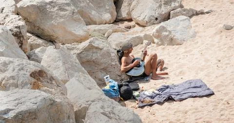Man bare-chested wearing flat checked cap sitting on beach playing guitar. Stock Photos