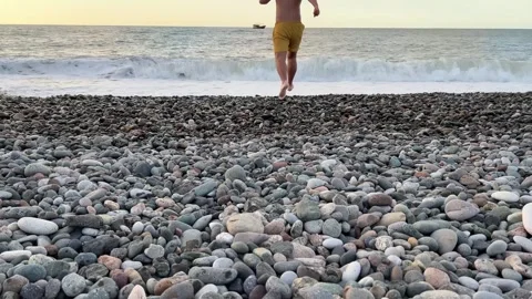 Man with bare torso in yellow shorts and transparent shoes runs into sea Stock Footage 255388472