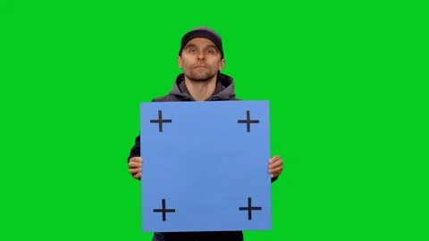 Man in baseball cap holding a blank board with tracking points on green screen 스톡 동영상 130794411