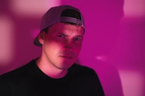 Man in baseball cap looking at camera under neon pink light. Serious young .. Stock Photos