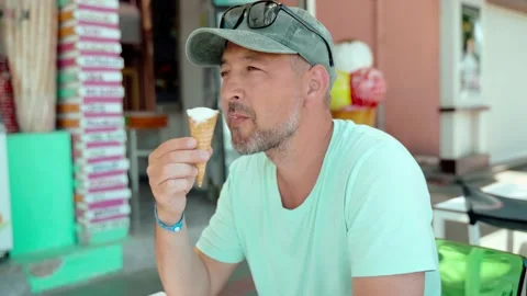 A man in a baseball cap sits and eats ice cream. A tourist eats an ice-cream in Stock Footage 165946963
