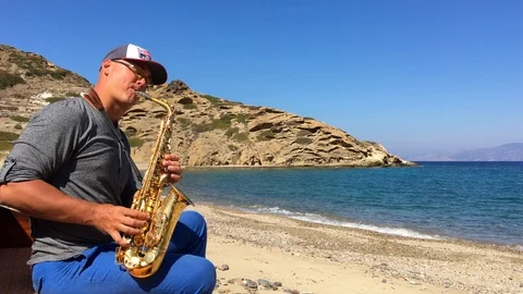 A man in a baseball cap sits on a sofa by the sea and plays the saxophone Stock Footage 86561866