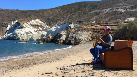 A man in a baseball cap sits on a sofa by the sea and plays the saxophone Stock Footage 86562003