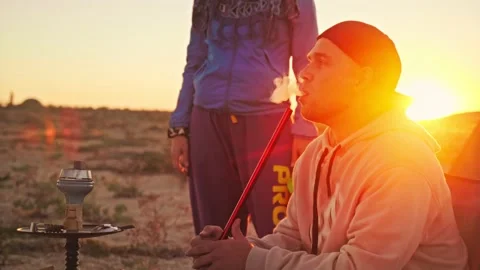 Man in baseball cap smokes a hookah on sandy beach against the setting sun Stock Footage 197332042