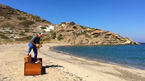 A man in a baseball cap stand on a sofa by the sea and plays the saxophone Stock Footage 86561915