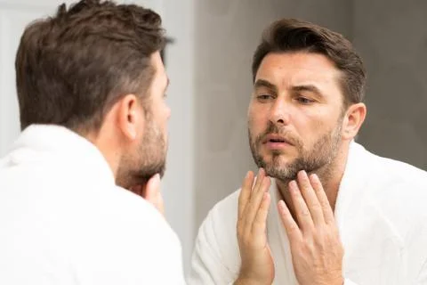 A man in a bathrobe in the bathroom looking at his wrinkles and pimples on face Stock Photos