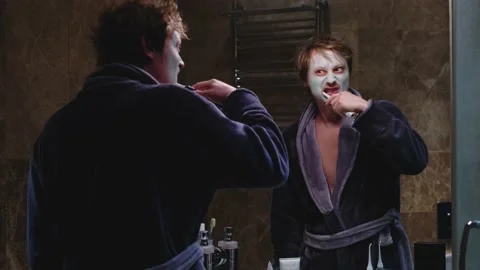 A man in a bathrobe brushes his teeth while looking in the mirror in the Vídeo Stock 194325880