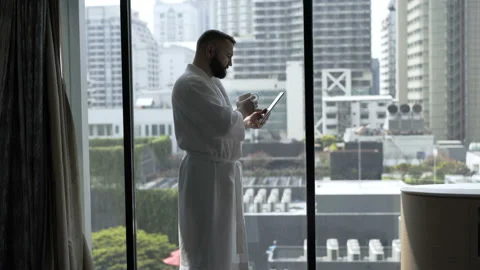 Man in bathrobe drinks coffee and uses tablet in his hotel room  Stock Footage 88437677