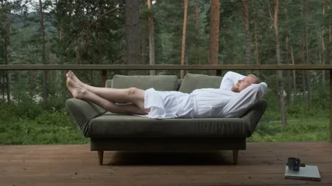 A man in a bathrobe lies on a sofa on a terrace in the forest Stock Footage 154278153